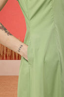 Close-up of a person wearing a light sage green dress, showing the side seam and a hand pocket with a tattooed arm tucked inside, against a coral and beige background.