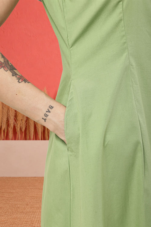 Close-up of a person wearing a light sage green dress, showing the side seam and a hand pocket with a tattooed arm tucked inside, against a coral and beige background.
