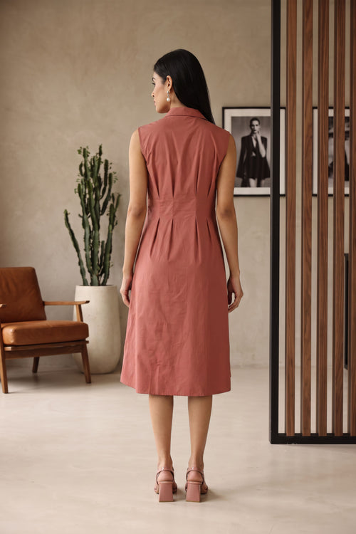 A woman stands with her back to the camera wearing a sleeveless dusty rose midi dress with a gathered waist, set in a modern interior room with a brown leather chair, potted cactus, and wooden slat screen.