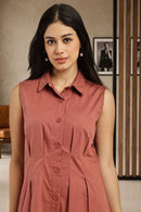 A woman wearing a sleeveless collared button-front dress in a dusty rose or terracotta color, standing indoors against a neutral wall with framed pictures and a slatted wooden screen.
