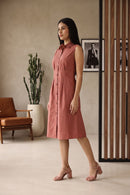 A woman models a sleeveless, button-front midi dress in a dusty rose pink color, standing in a modern indoor setting with a brown leather chair, a potted cactus, and a wooden slatted screen.