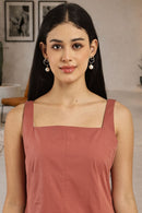 A woman with long dark hair wearing a dusty rose square-neck top and silver hoop earrings with white pearl drops, standing in a modern indoor setting.