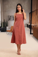 A woman modeling a sleeveless, square-neck midi dress in a dusty rose or terracotta color, standing in a modern indoor room with neutral-toned walls and furniture.