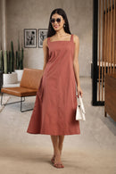 A woman wearing a dusty rose midi dress with a square neckline and thin shoulder straps, standing in a modern indoor setting with a brown leather chair, potted cacti, and a wooden slatted screen.