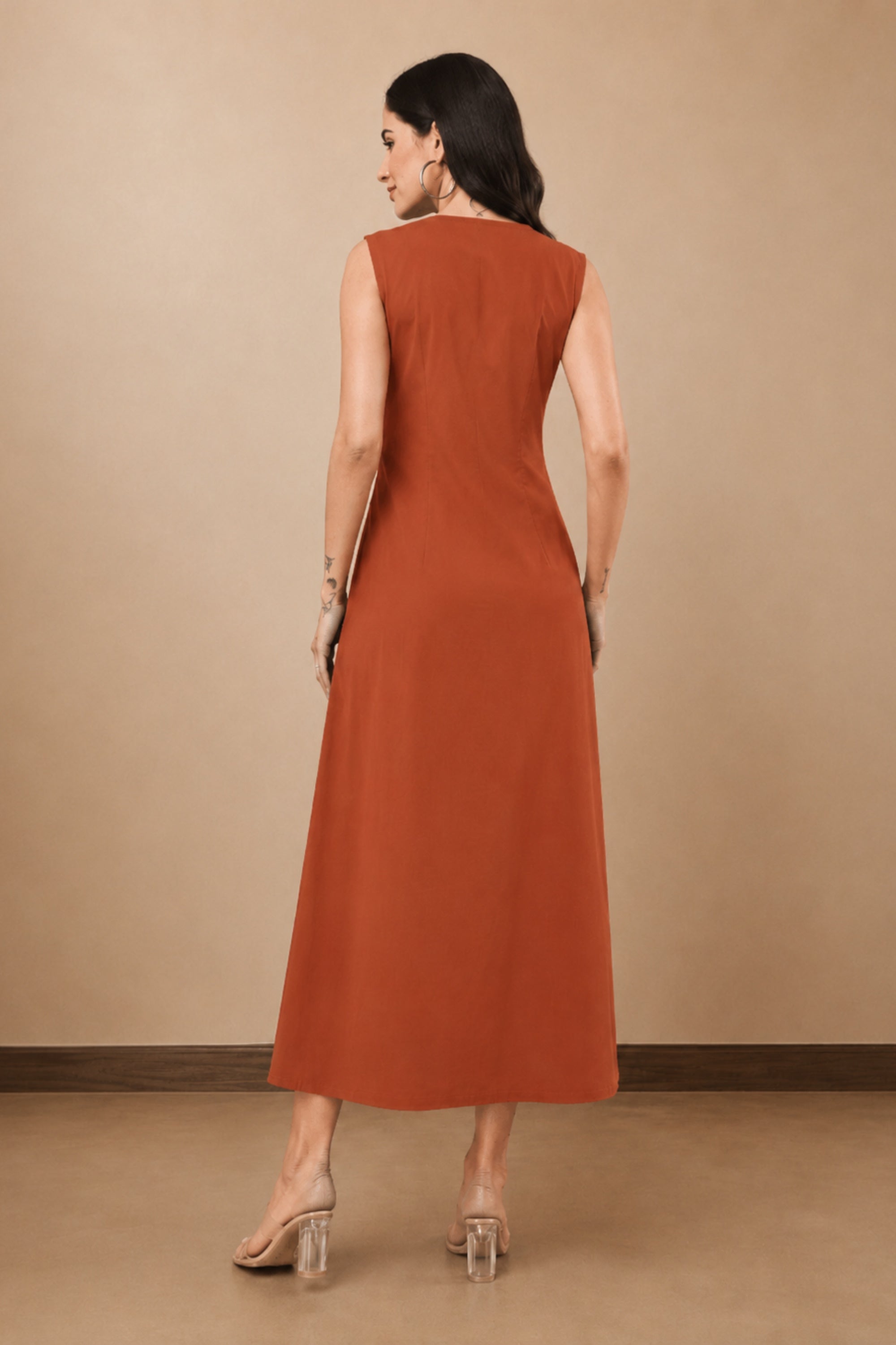 A woman wearing a sleeveless rust-brown midi dress, shown from the back against a plain beige studio backdrop.