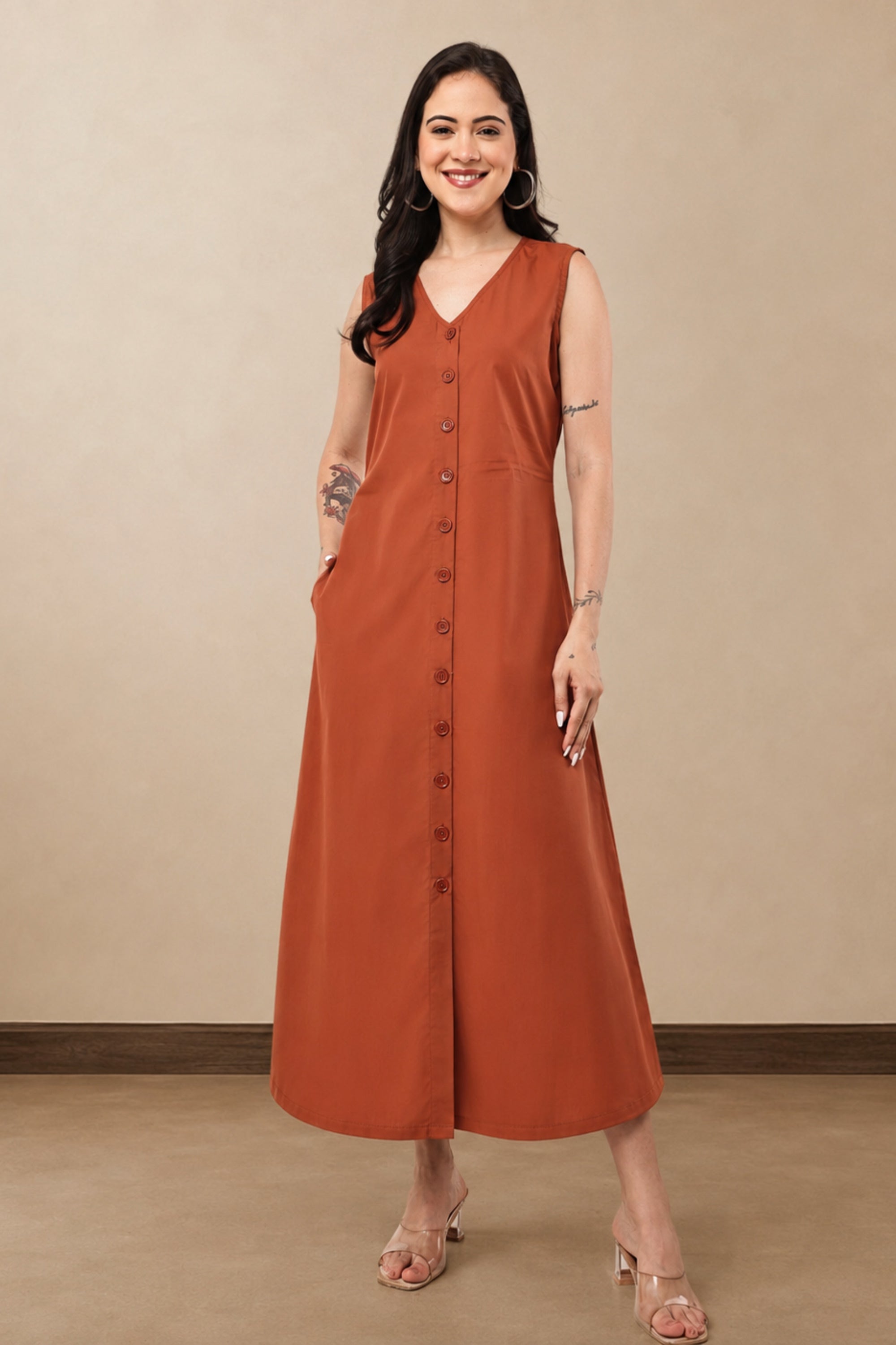 A woman modeling a sleeveless rust-orange midi dress with a V-neck, front button closure, and side pockets, standing against a plain beige studio wall.