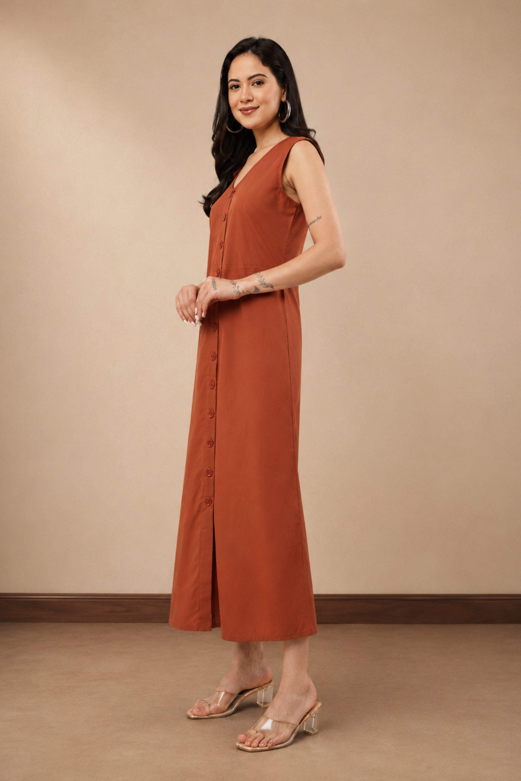 A woman modeling a sleeveless rust-orange maxi dress with a button-front detail, standing against a plain beige studio wall.