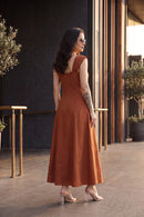 A woman wearing a rust-orange sleeveless maxi dress with a scoop neck and back, styled with clear high-heeled sandals, standing in an outdoor patio setting.
