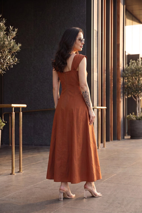 A woman wearing a rust-orange sleeveless maxi dress with a scoop neck and back, styled with clear high-heeled sandals, standing in an outdoor patio setting.