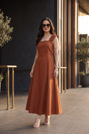 A woman modeling a rust-orange sleeveless midi dress with a square neckline and a flowing skirt, standing in an outdoor patio setting with a dark textured wall and vertical wooden slats.