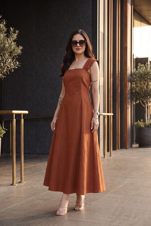 A woman modeling a rust-orange sleeveless midi dress with a square neckline and a flowing skirt, standing in an outdoor patio setting with a dark textured wall and vertical wooden slats.