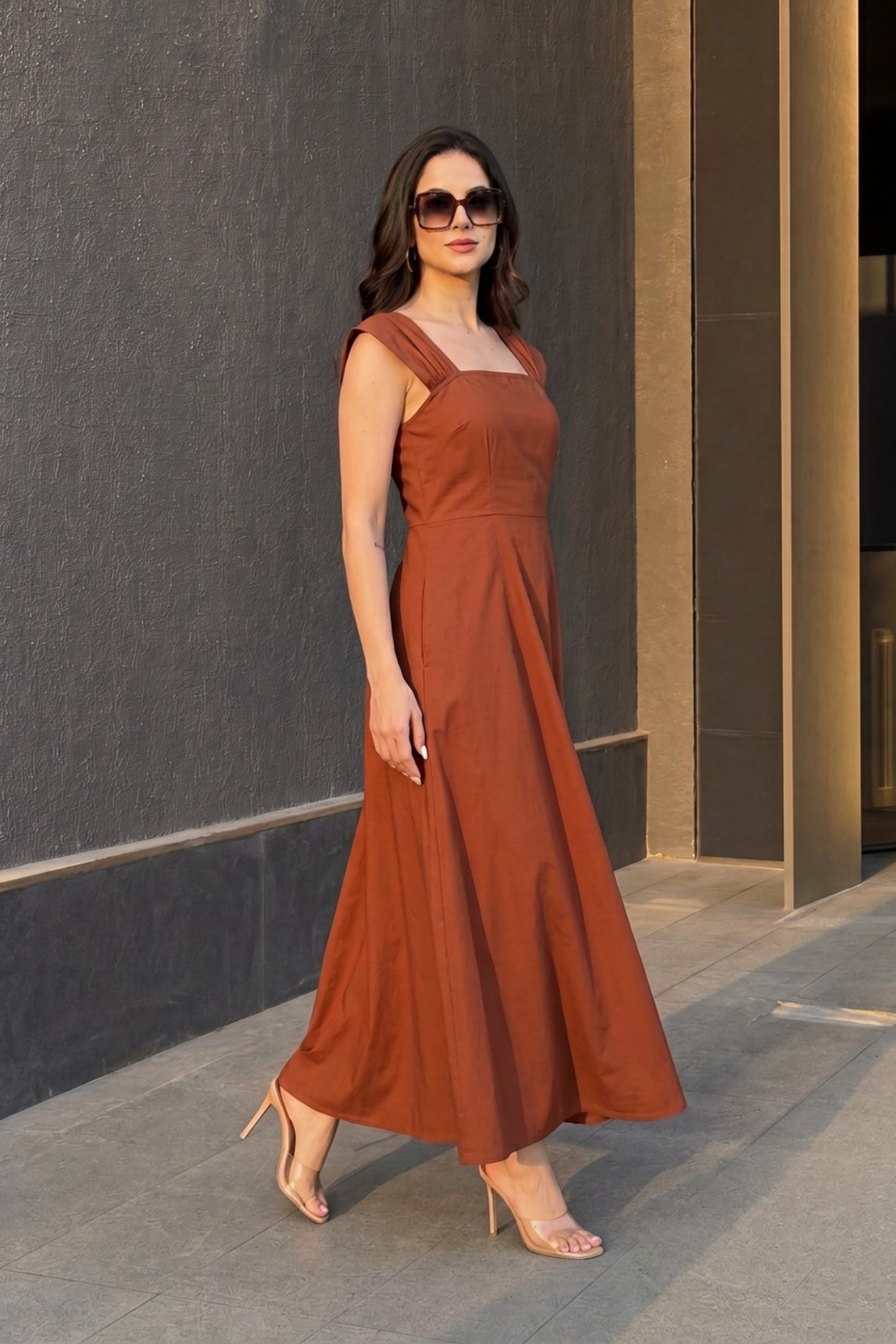 A woman wearing a rust-orange sleeveless maxi dress with a square neckline and a flowing skirt, styled with nude high heels and sunglasses, standing outdoors against a dark gray textured wall.