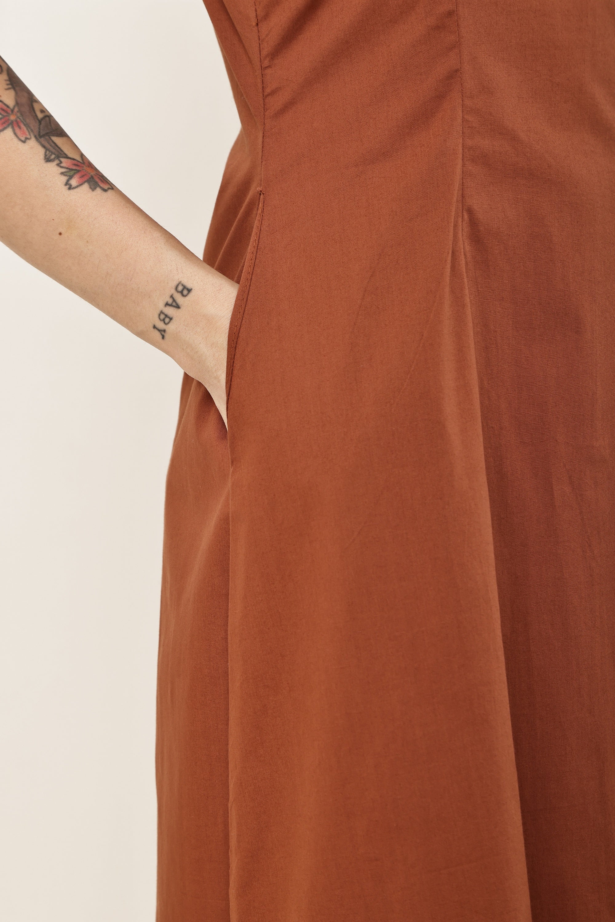Close-up of a terracotta rust-colored dress with a side pocket, worn by a model whose tattooed arm with the word 'BAPT' is visible on the left, against a plain off-white background.