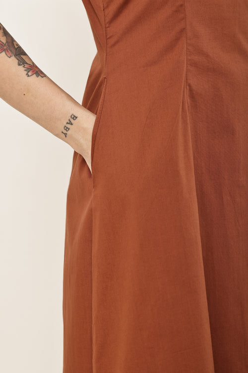 Close-up of a terracotta rust-colored dress with a side pocket, worn by a model whose tattooed arm with the word 'BAPT' is visible on the left, against a plain off-white background.