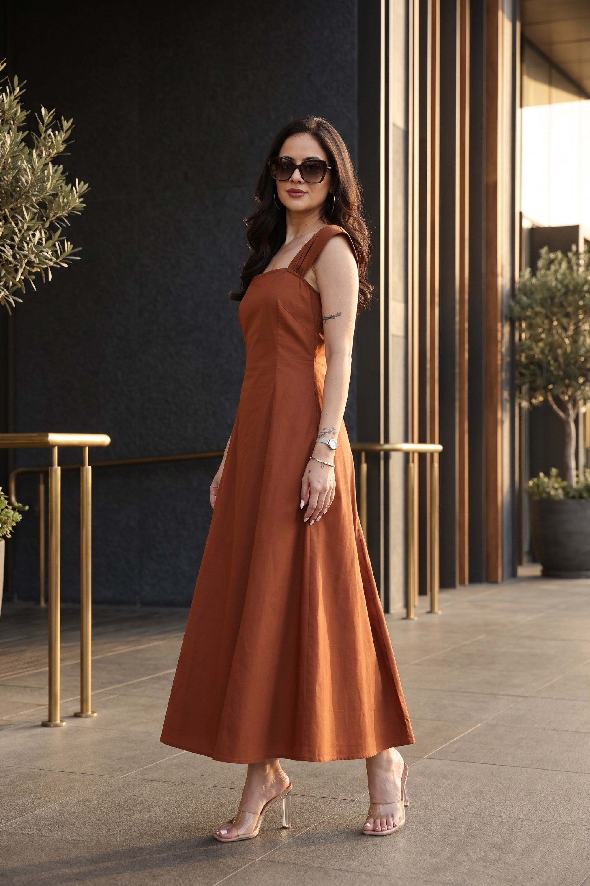 A woman wearing a rust-orange sleeveless midi dress with a V-neckline and A-line silhouette, styled with nude high-heeled sandals and sunglasses, standing in a modern outdoor corridor.
