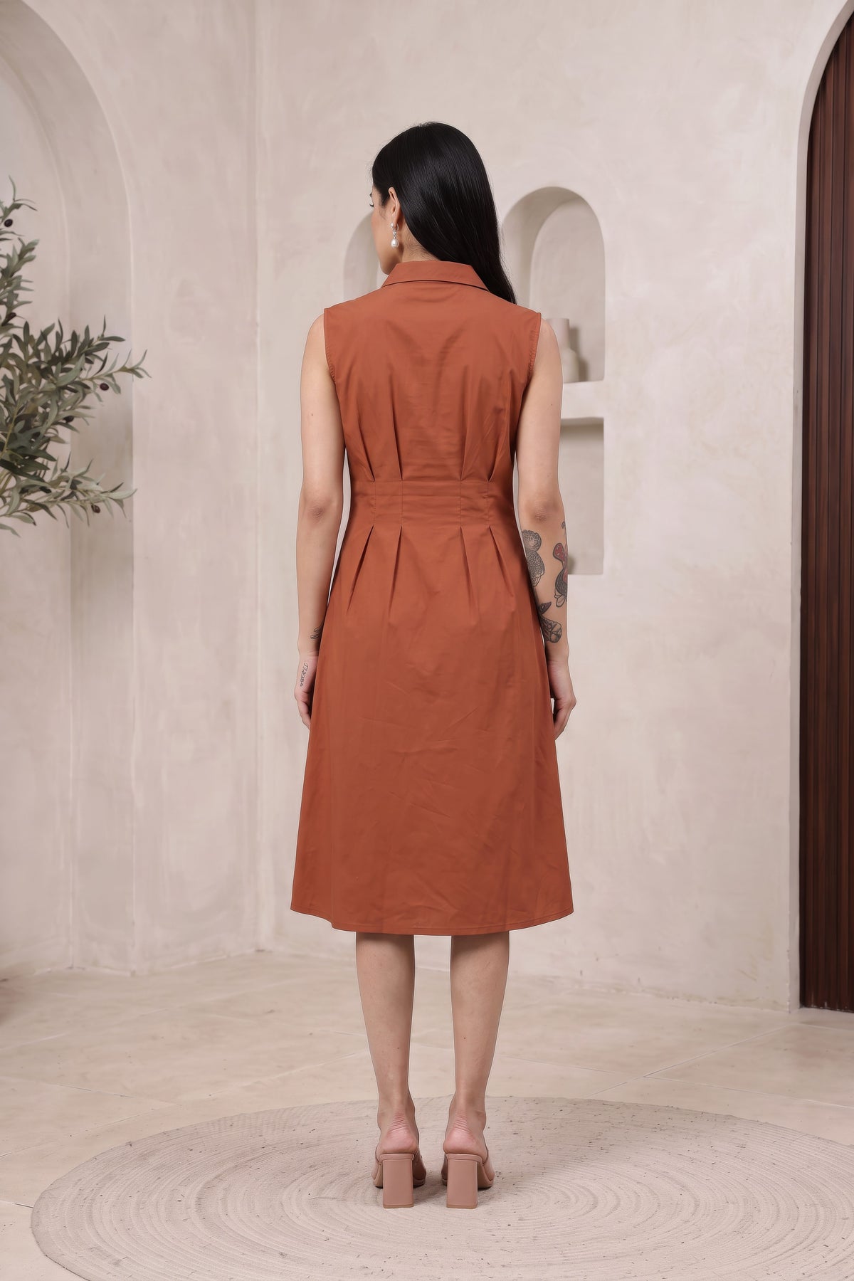 A woman viewed from behind wearing a sleeveless, collared rust-orange dress with a gathered waist, standing in a minimalist cream-colored room with arched alcoves.
