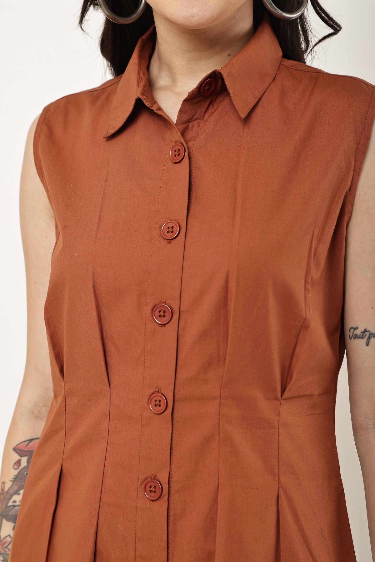 A close-up of a sleeveless collared button-front top or dress in a warm rust orange color, worn by a model. The garment features a pointed collar, a vertical button placket with matching buttons, and a slightly gathered silhouette.