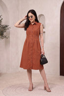 A woman models a sleeveless, button-front midi dress in a warm rust or terracotta color. The dress has a collared neckline and a flared skirt, shown in a styled indoor setting with a beige textured wall, arches, and a small black handbag.
