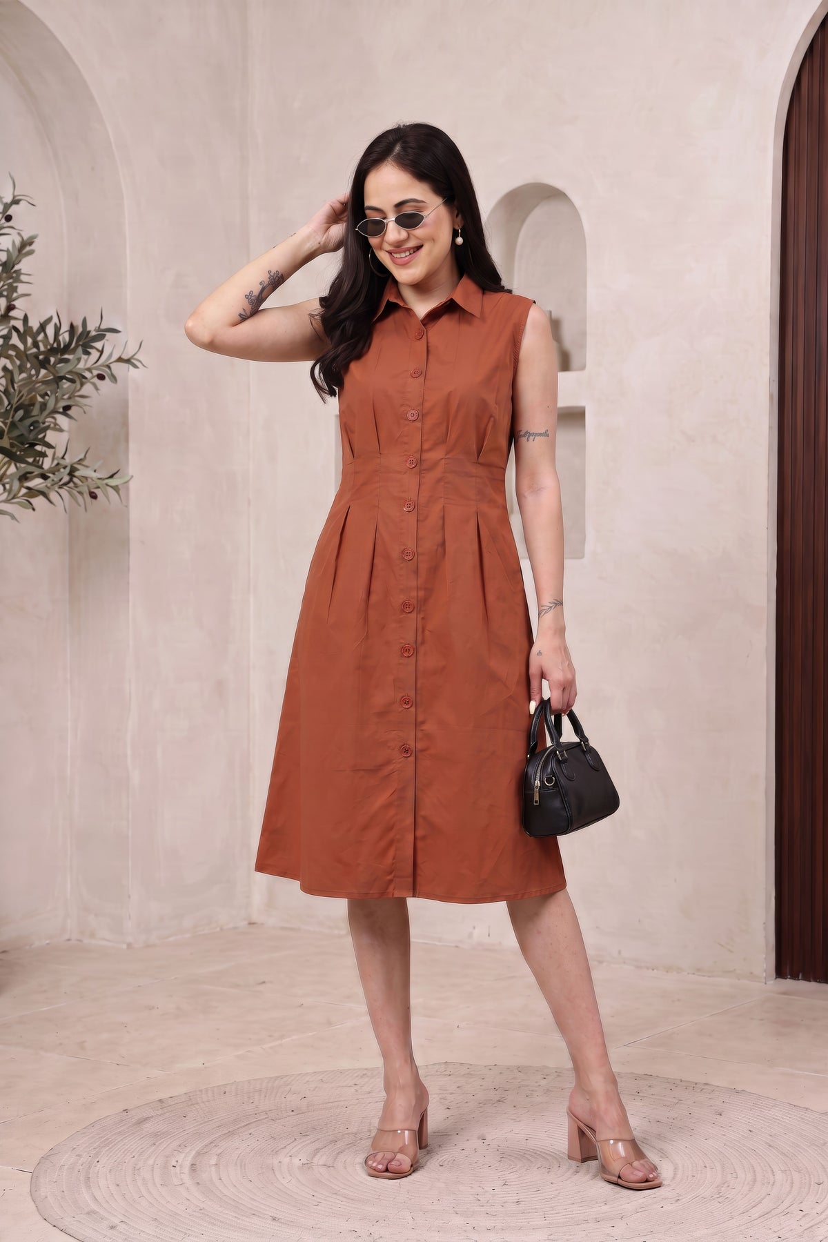 A woman models a sleeveless, button-front midi dress in a warm rust or terracotta color. The dress has a collared neckline and a flared skirt, shown in a styled indoor setting with a beige textured wall, arches, and a small black handbag.