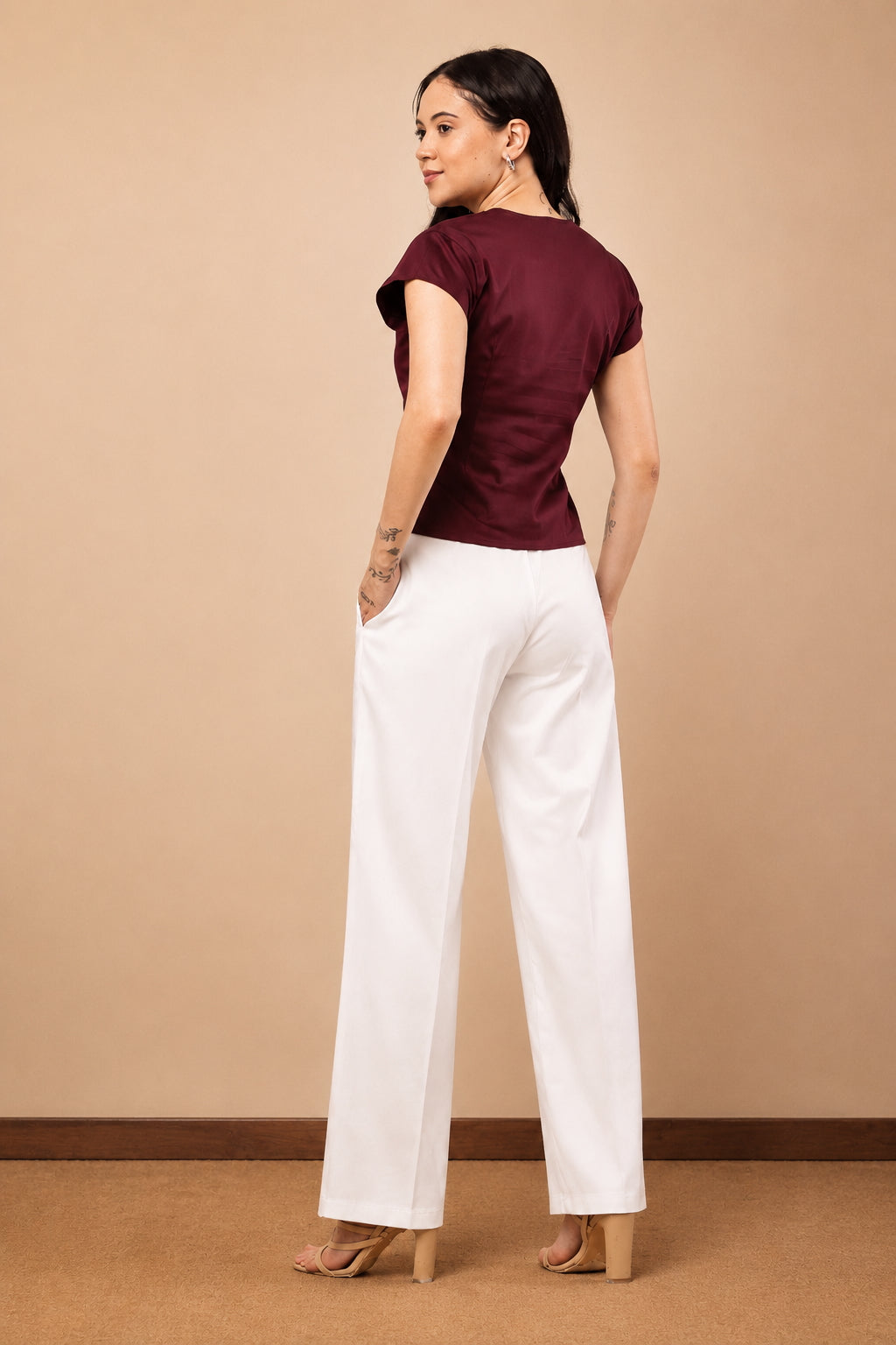 A woman stands in a studio wearing a short-sleeve burgundy top and white wide-leg pants, photographed from the back against a plain beige backdrop.