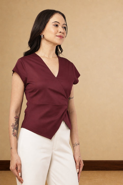A woman modeling a short-sleeve wrap-style blouse in deep burgundy with white pants, standing against a warm beige studio wall.