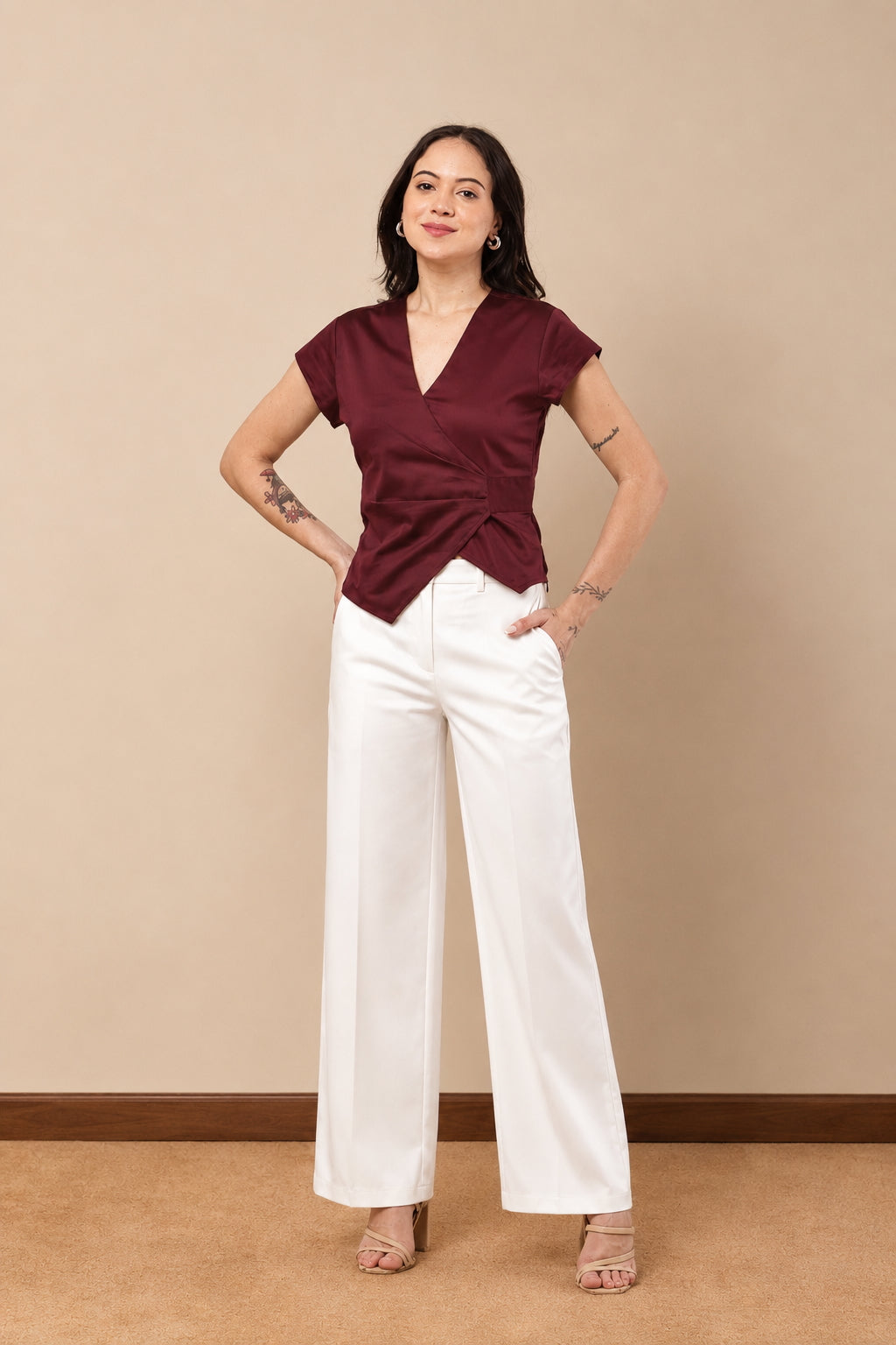 A woman modeling a short-sleeve wrap-style top in deep burgundy with a twisted front detail, paired with white wide-leg pants, standing against a beige studio wall.