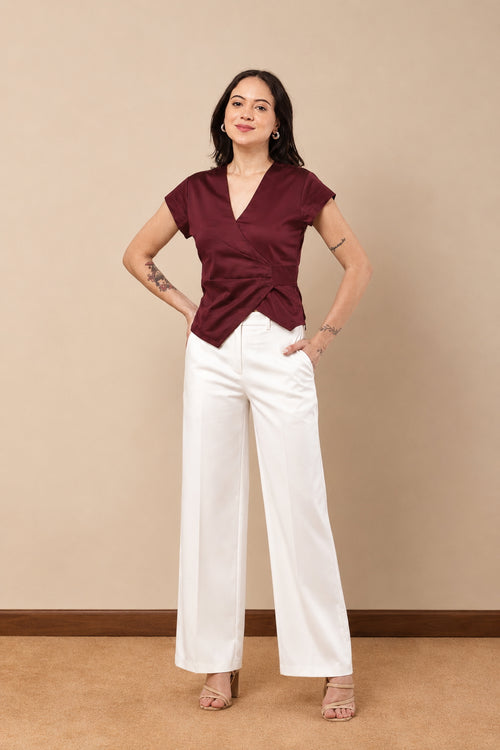 A woman modeling a short-sleeve wrap-style top in deep burgundy with a twisted front detail, paired with white wide-leg pants, standing against a beige studio wall.
