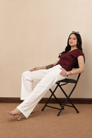 A woman modeling a short-sleeve wrap-style top in deep burgundy with a twisted front detail, paired with white wide-leg pants, sit on black chair a beige studio wall.