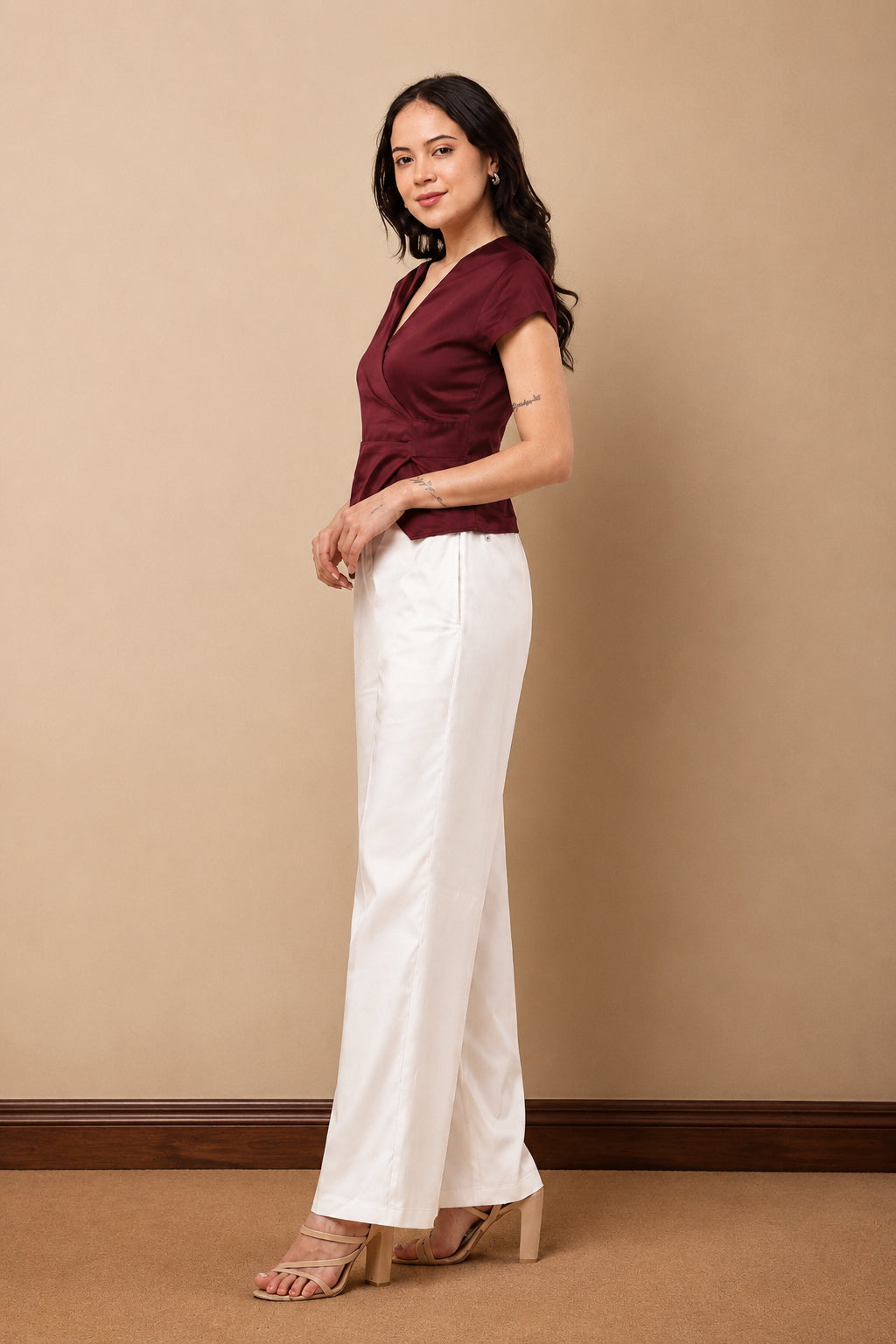 A woman modeling a burgundy short-sleeve wrap-style top and white wide-leg pants, standing against a plain beige studio wall.