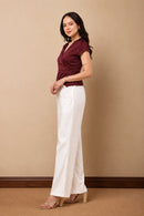 A woman modeling a burgundy short-sleeve wrap-style top and white wide-leg pants, standing against a plain beige studio wall.