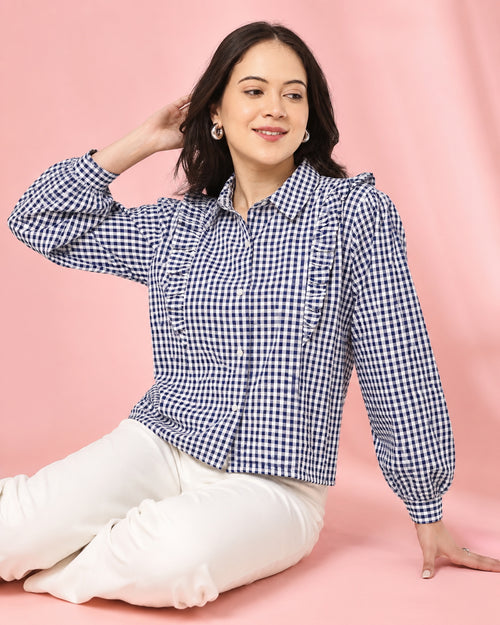 A woman wearing a navy and white gingham long-sleeve blouse with puff sleeves and white pants, posing against a solid pink studio backdrop.