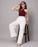 A woman posing on a wooden stool wearing a maroon halter crop top, white wide-leg trousers, and clear high-heel sandals against a solid gray studio background.